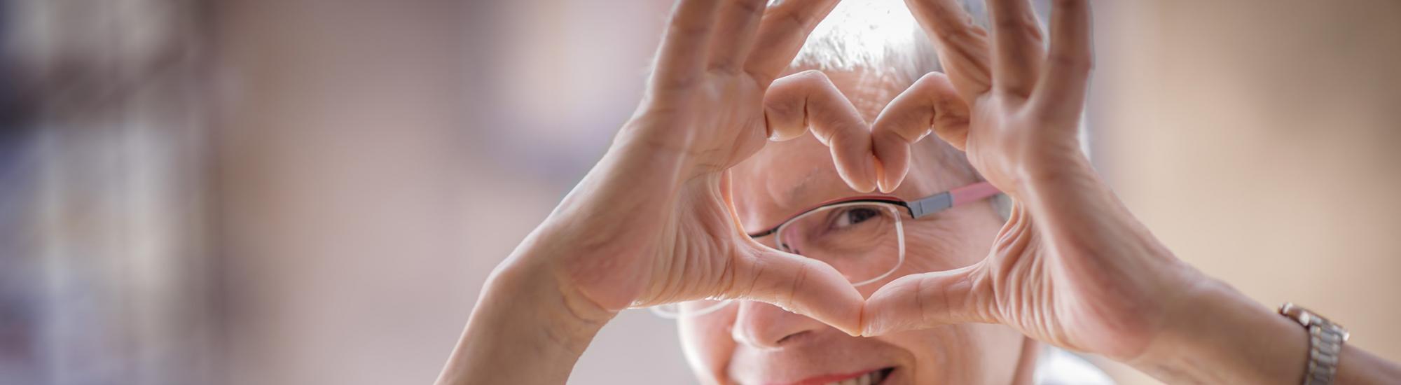 An older adult woman making a heart with her fingers.